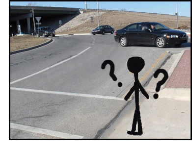 Photo shows a stick figure facing a crosswalk across two lanes.  Question marks are drawn around his head.  About 20 feet to our right, our street enters a roundabout (lanes go around a large circle).  Cars are coming from the other side of the circle and approaching the crosswalk.